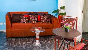 Een woonkamer met een blauwe muur, een plafondventilator, een bank met bloemenkussens, een glazen salontafel en een eettafel met stoelen.