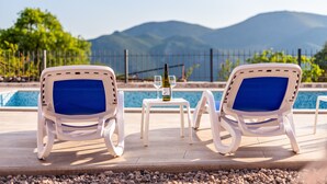 Zwei weiße Lounge-Stühle mit blauen Kissen, ein kleiner Tisch mit einer Weinflasche und zwei Gläsern, sowie ein Pool mit Blick auf eine Berglandschaft im Hintergrund.