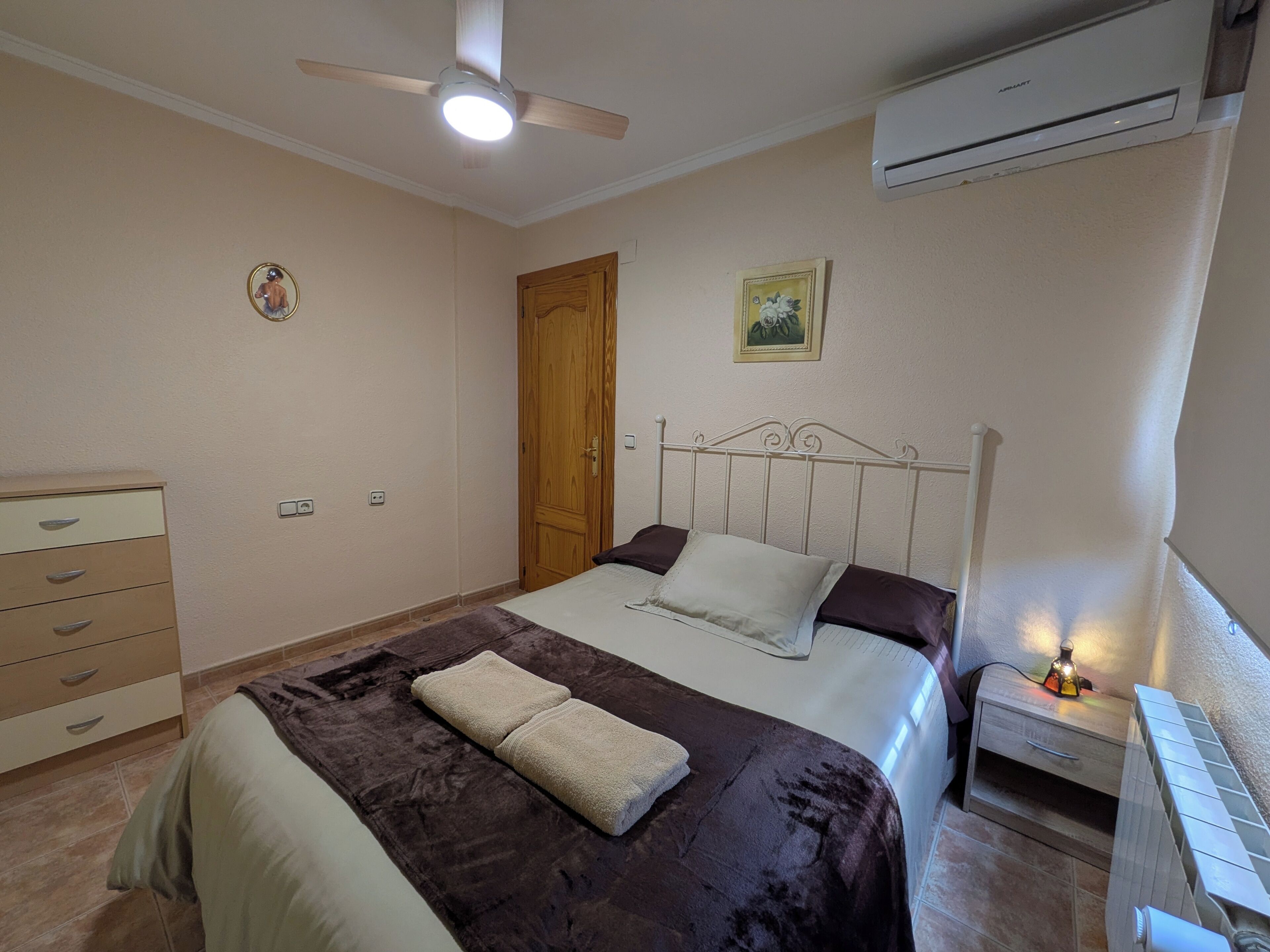 Une chambre à coucher comprenant un lit, une table de chevet, une commode, un ventilateur de plafond, un appareil de climatisation et une porte.