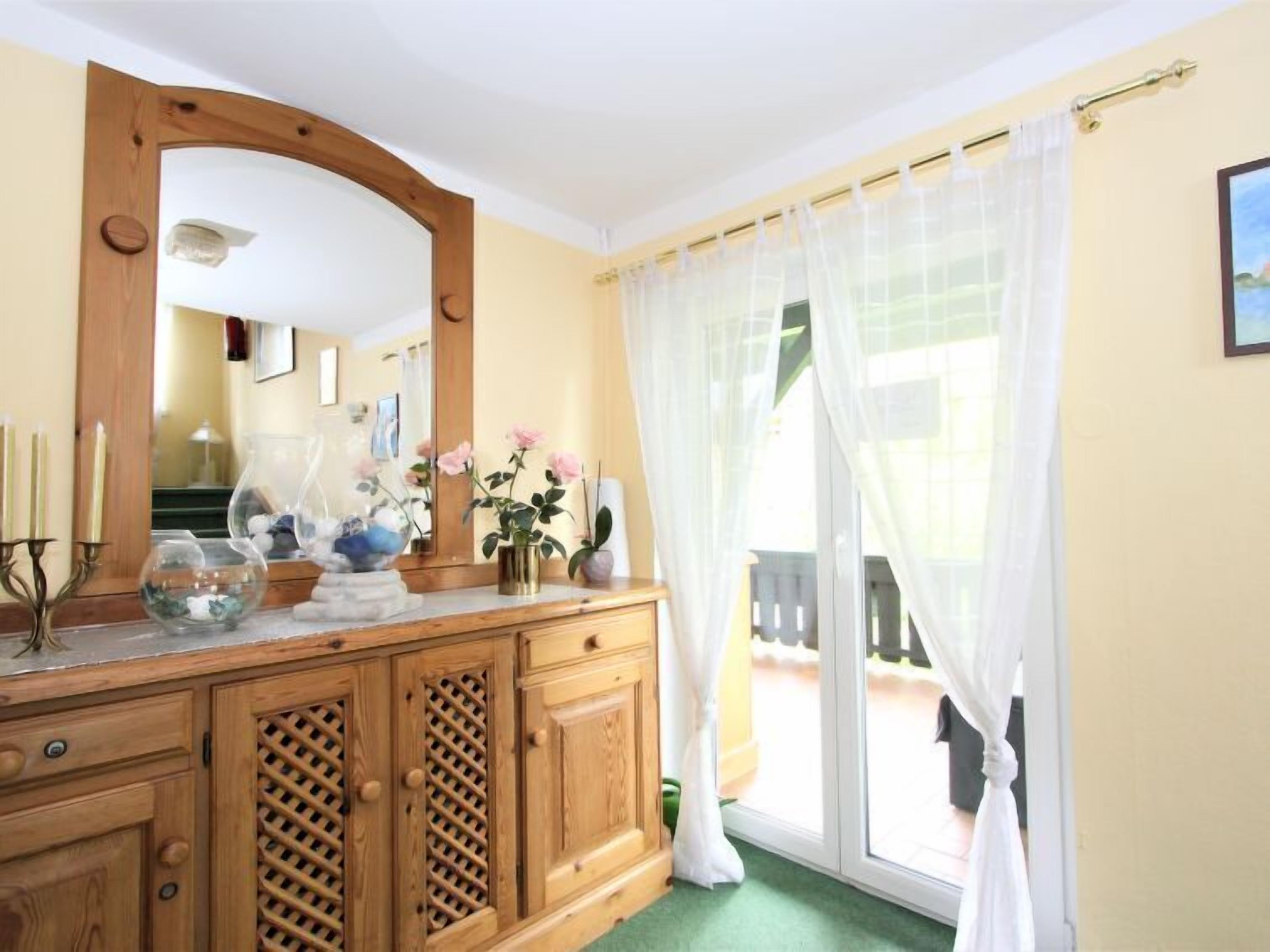 A room with a wooden dresser, a mirror, a window with sheer curtains, and a view of a balcony.