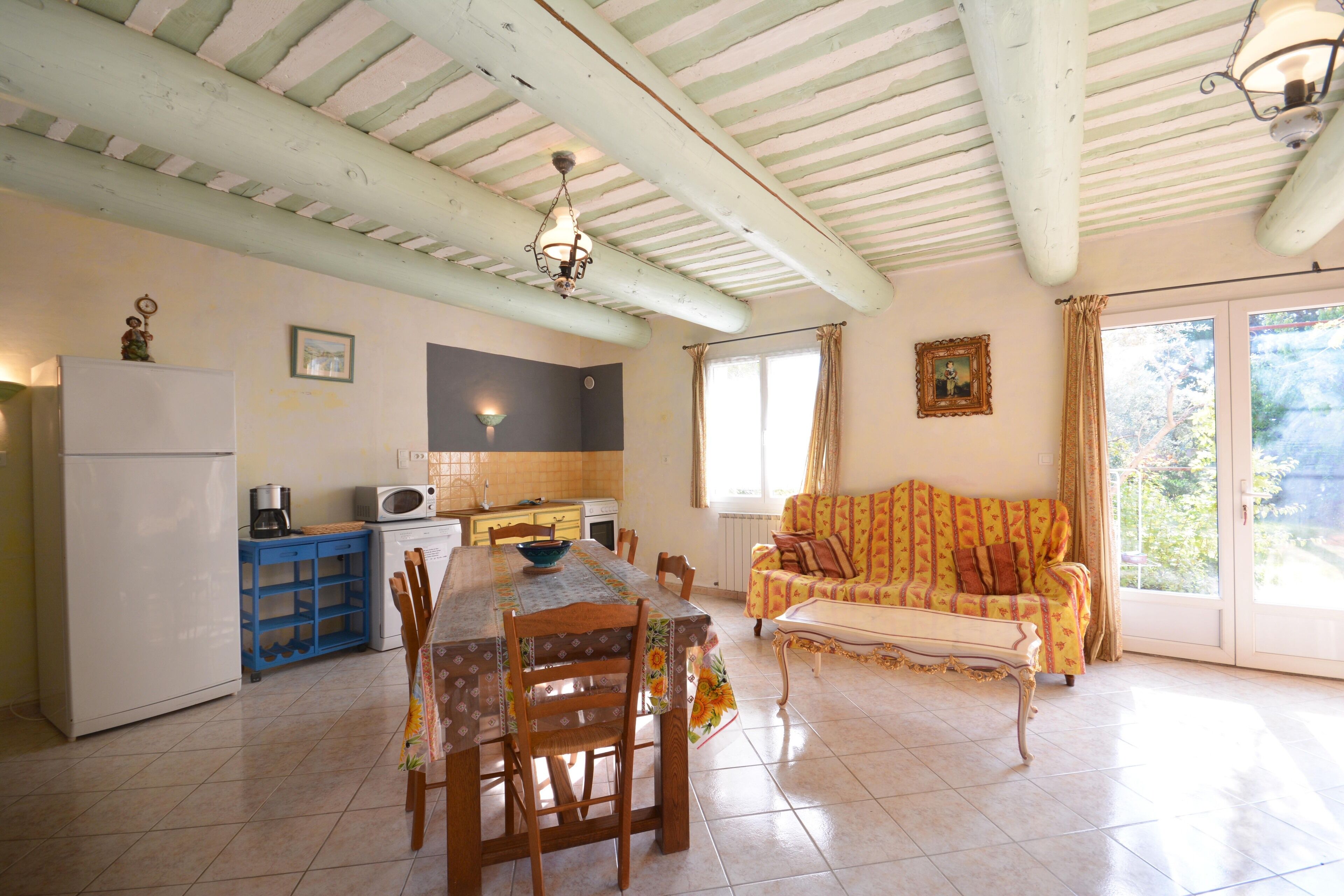 A kitchen with a dining table, chairs, and a sofa. There is a refrigerator, microwave, and a window with curtains.