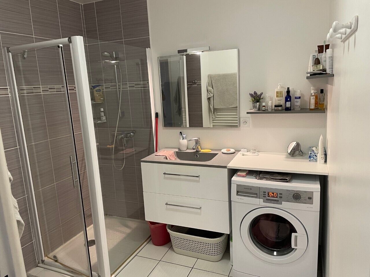 A modern bathroom with a glass shower enclosure, a built-in washing machine, and a vanity with a mirror and shelves.
