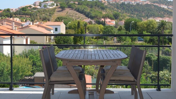 A balcony with a round table and two chairs, overlooking a hillside with houses and greenery.