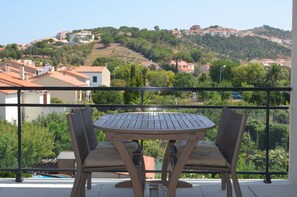 A balcony with a round table and two chairs, overlooking a hillside with houses and greenery.
