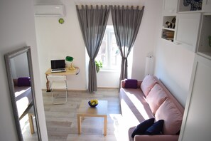 Un salon avec un canapé rose, une table basse en bois, un ordinateur portable posé sur un bureau et une fenêtre avec des rideaux.
