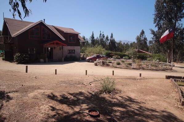 A rustic building with a red roof and wooden siding, a fenced area with a red car, and a flagpole with a flag. - Casa en Parcela (Zapallar)