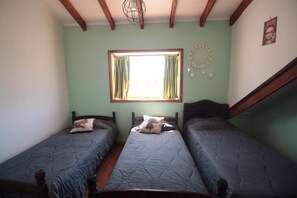 A room with two single beds, a window with green curtains, a dreamcatcher on the wall, and a wooden ceiling beam. - Casa en Parcela (Zapallar)