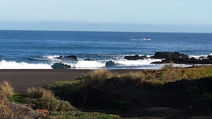 Une plage où les vagues se brisent sur le rivage, un bateau à l’horizon et des affleurements rocheux.
