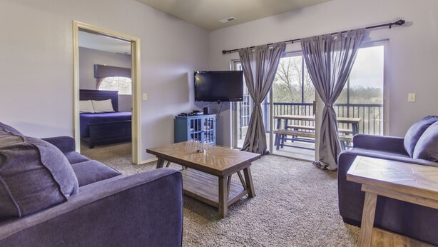 A living room with a sofa, a table, a TV, and a balcony with curtains. - Stonebridge Condo and Golf Resort (Branson West)