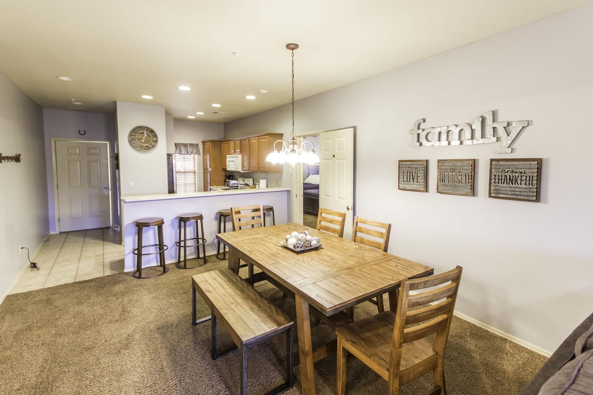A dining area with a wooden table, benches, and chairs. There is a kitchen in the background with wooden cabinets and a countertop.