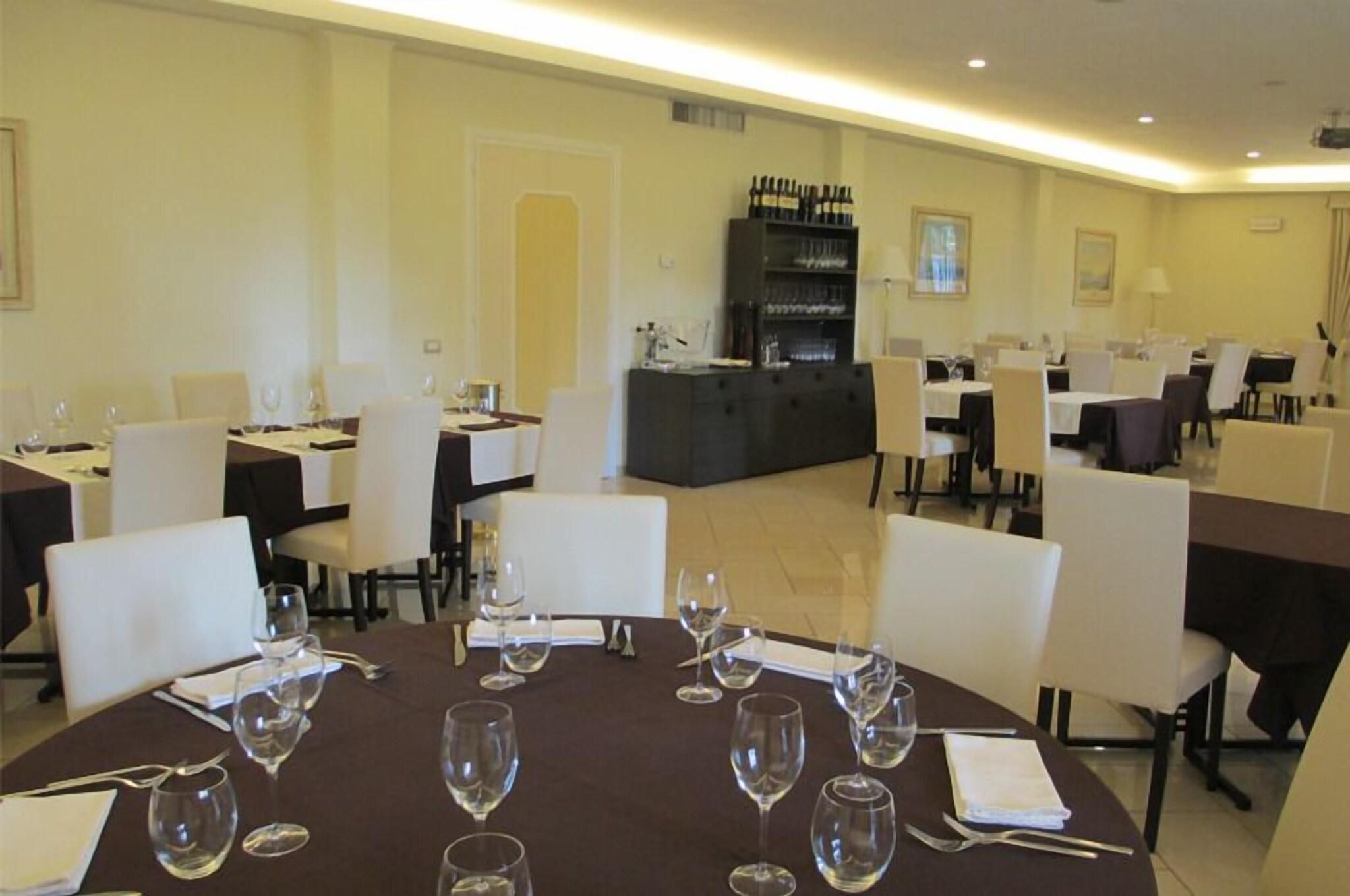 Uma sala de jantar com mesas redondas, cadeiras brancas e copos de vinho. Há também um bar com garrafas e copos.