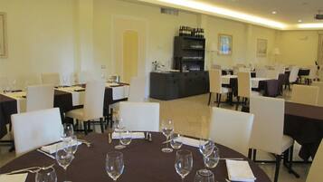 Uma sala de jantar com mesas redondas, cadeiras brancas e copos de vinho. Há também um bar com garrafas e copos.