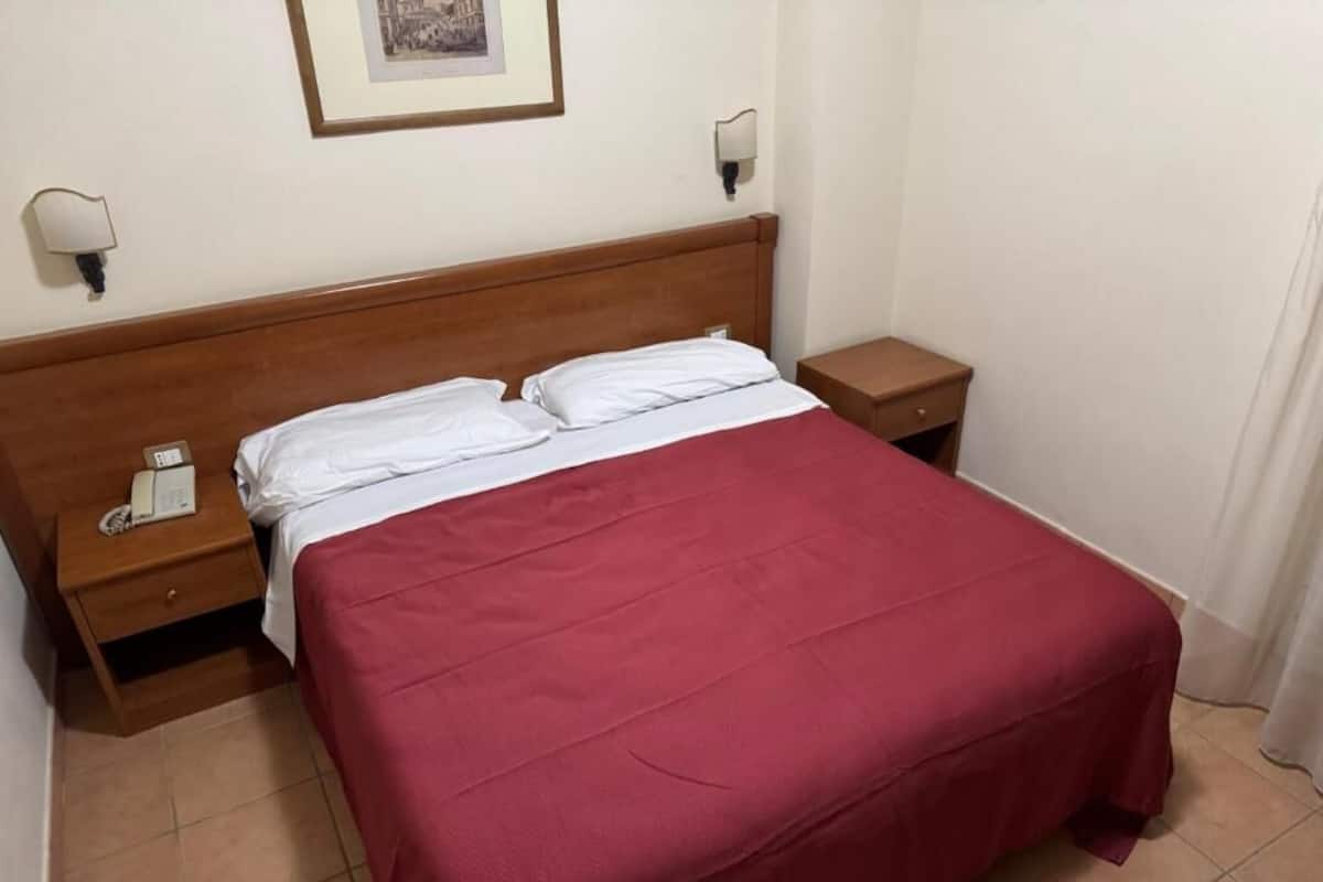 A single bed with a wooden headboard, a red bedspread, two bedside tables with lamps, and a framed picture on the wall.