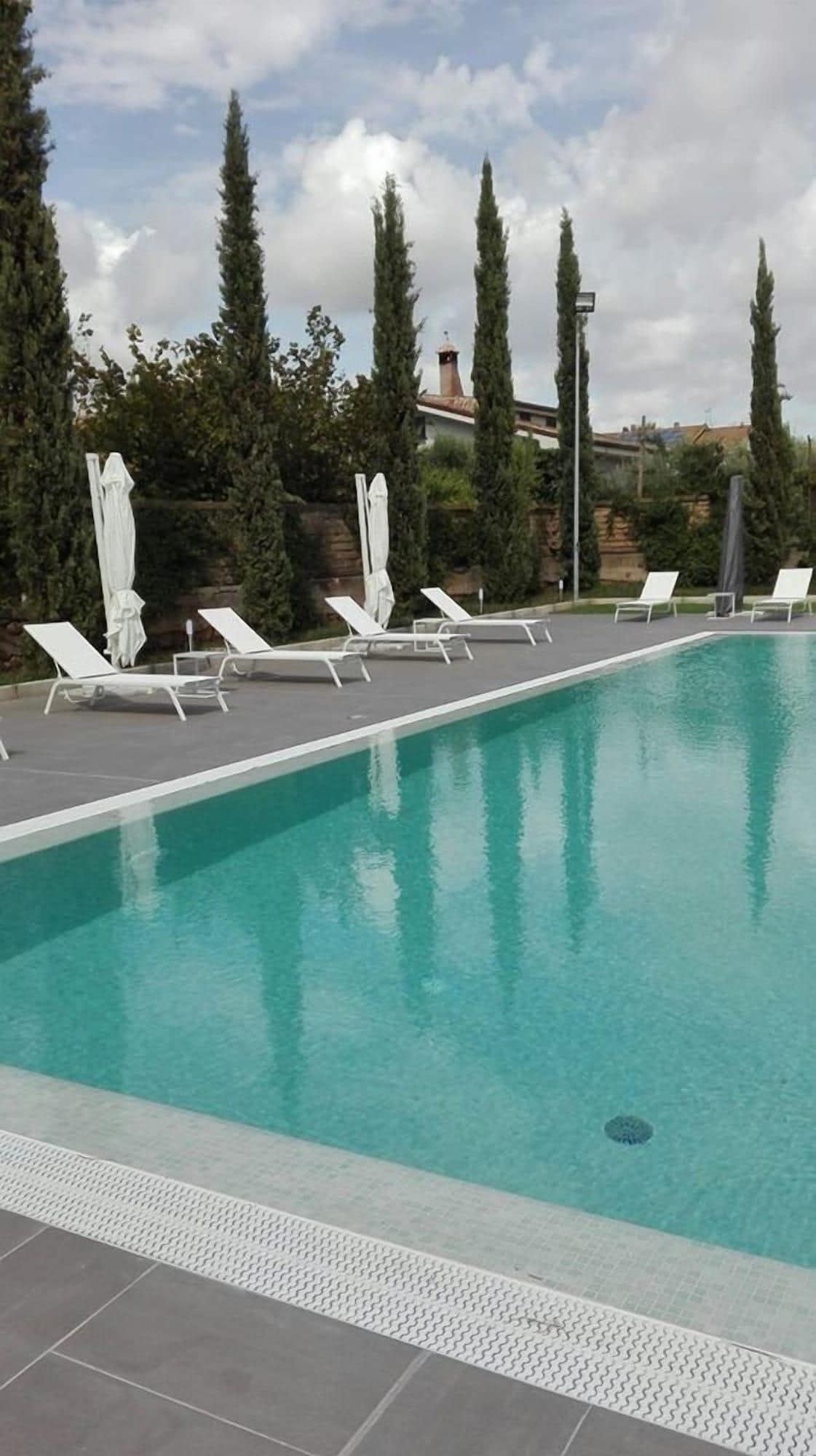 Una piscina rectangular amb gandules i ombrel·les blanques, envoltada d'altes palmeres i un cel nuvolós.