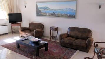 Uma sala de estar com dois sofás de couro marrom, uma mesa de centro, uma televisão sobre um móvel, uma pequena mesa lateral e uma pintura representando uma paisagem costeira.