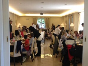 A hotel dining area with multiple tables and chairs, a central serving area, and a view of the outdoors through large windows. - Villamaria Park Hotel (Albano Laziale)