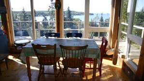 A dining area with a table and chairs, large windows with a view of a lake and forest, and wooden flooring. - Relaxing ocean front property with picturesque harbor views (Addison)