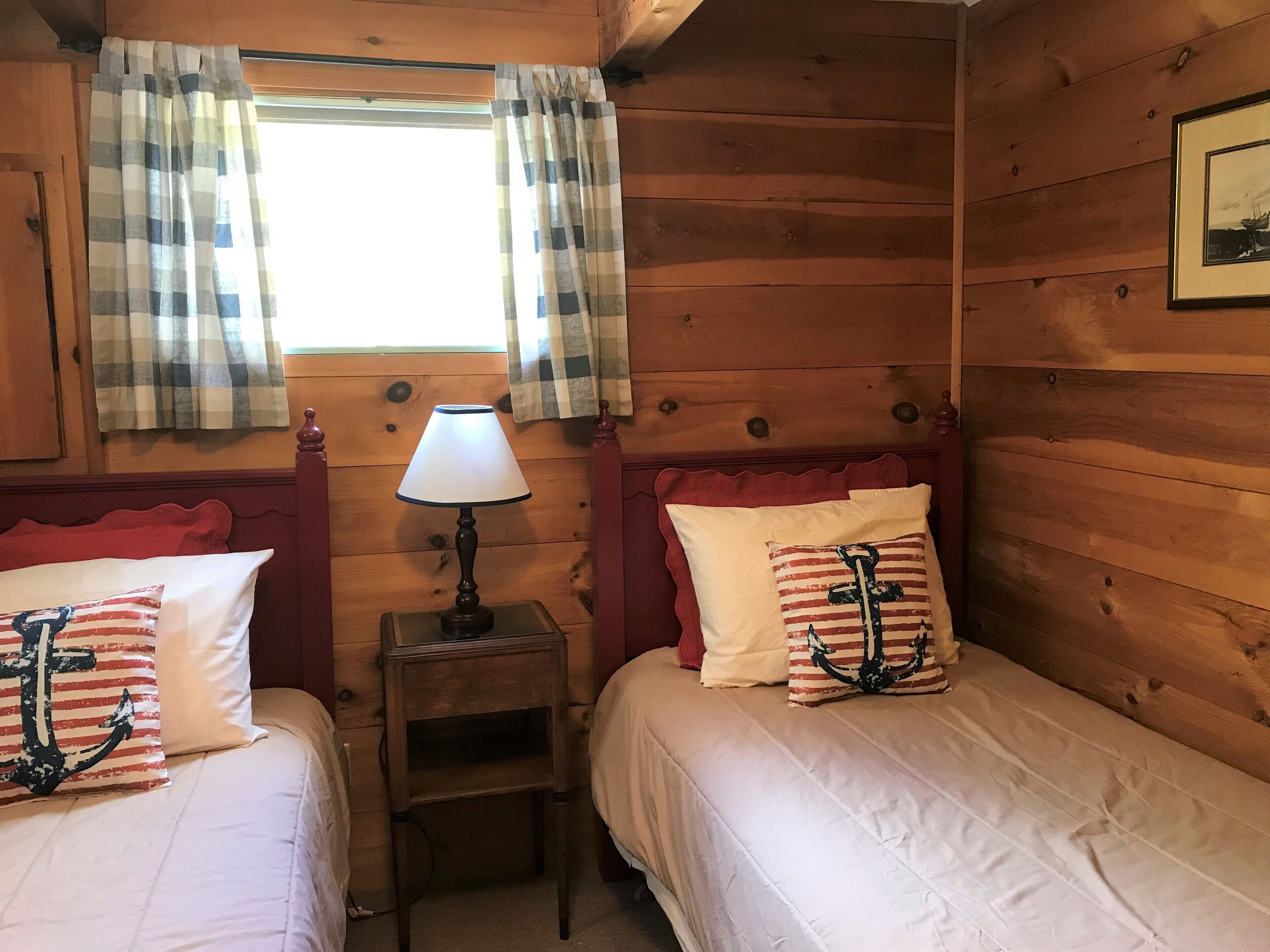 Una habitación tipo cabaña con dos camas, una mesita de noche con lámpara, cortinas escocesas y paredes de madera.