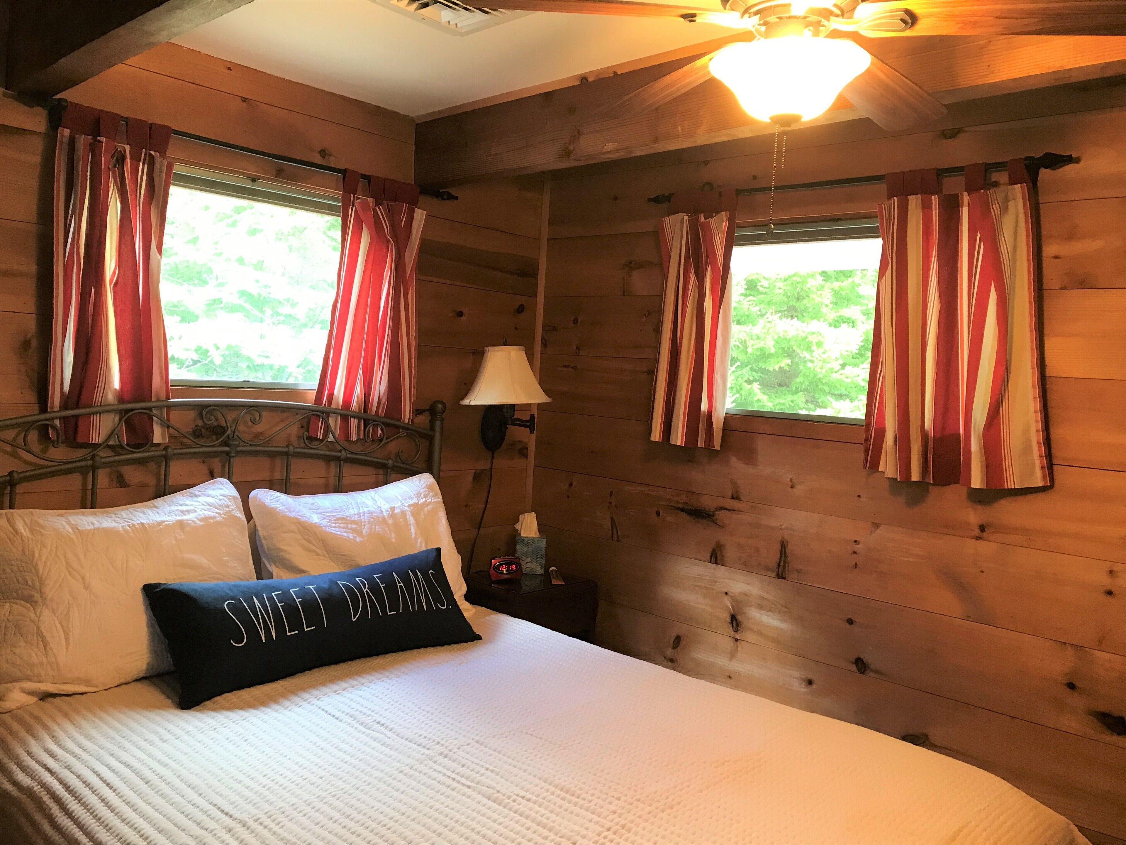 A wooden cabin bedroom with a bed, curtains, a lamp, and a clock.