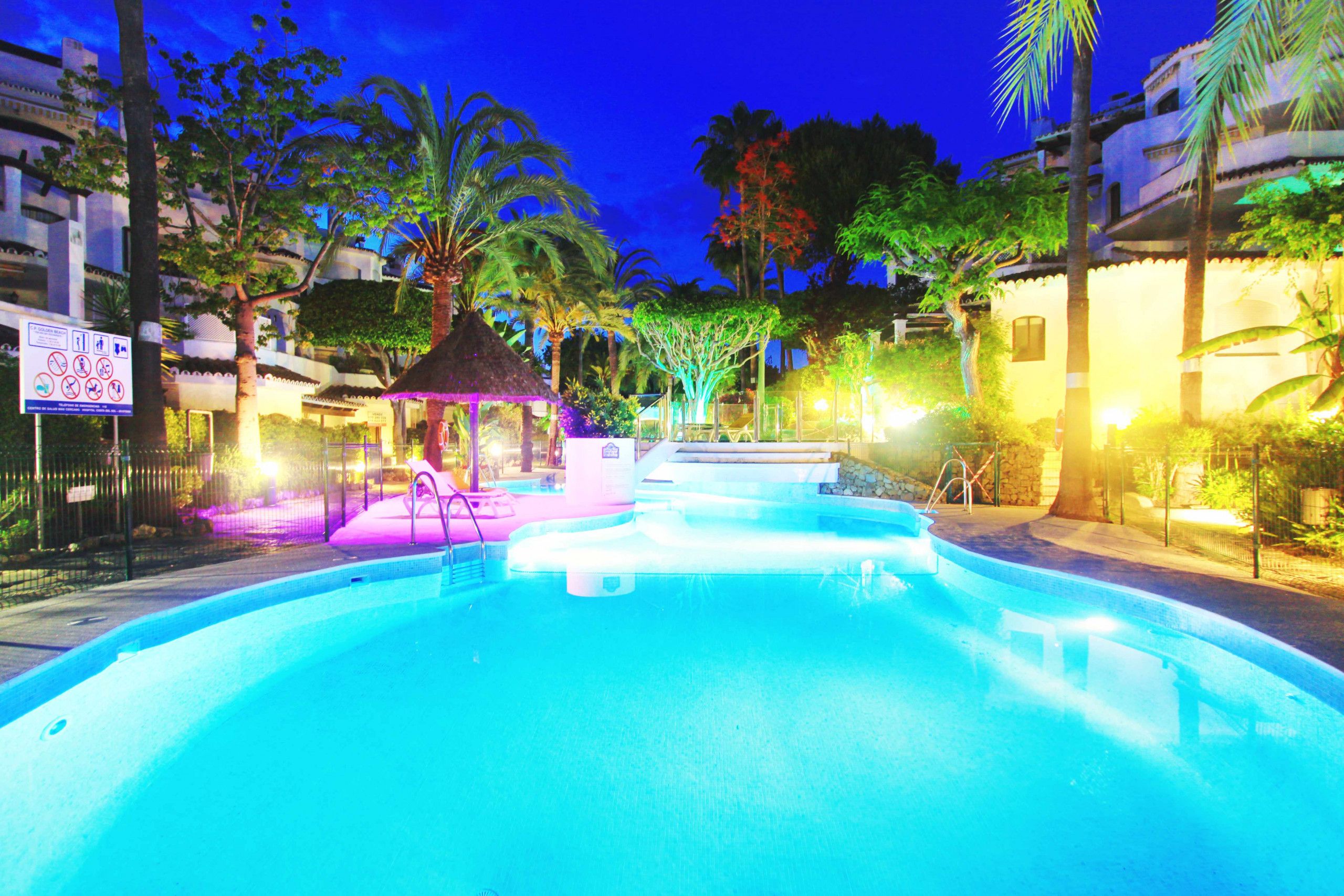 A swimming pool with a thatched-roof gazebo, surrounded by palm trees and a fence.