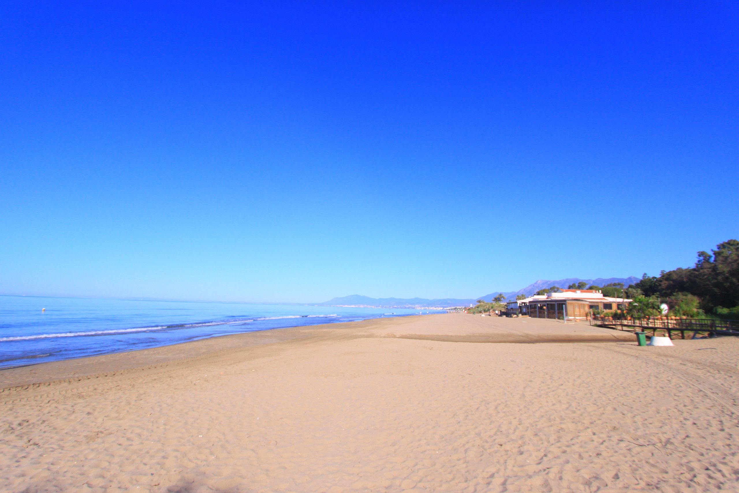 Ein weiter Sandstrand, strahlend blauer Himmel, einige Gebäude in der Ferne und ein Bergkamm im Hintergrund.