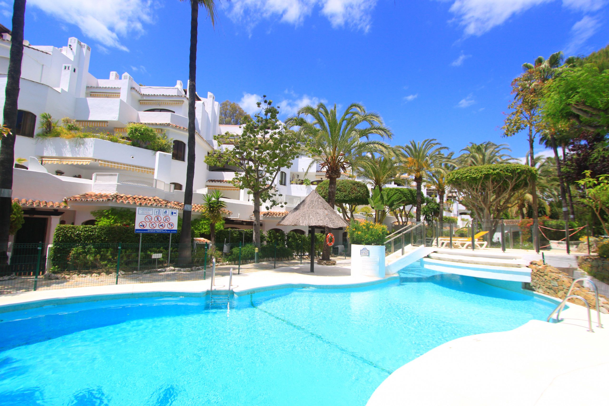 A swimming pool area with a thatched-roof structure, palm trees, and white buildings with balconies.