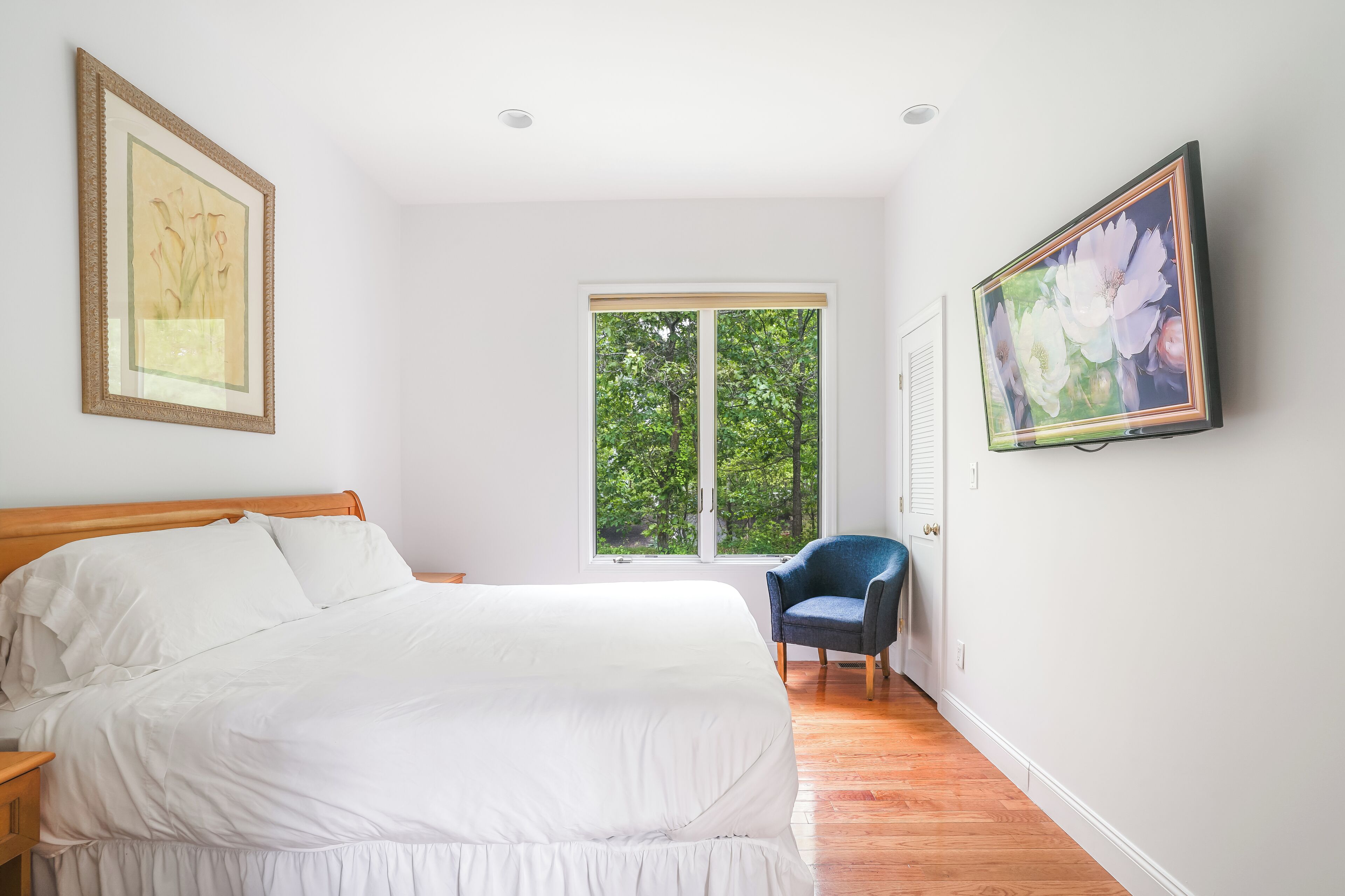 A bedroom with a bed, a chair, a window with a view of trees, and two framed pictures on the wall.