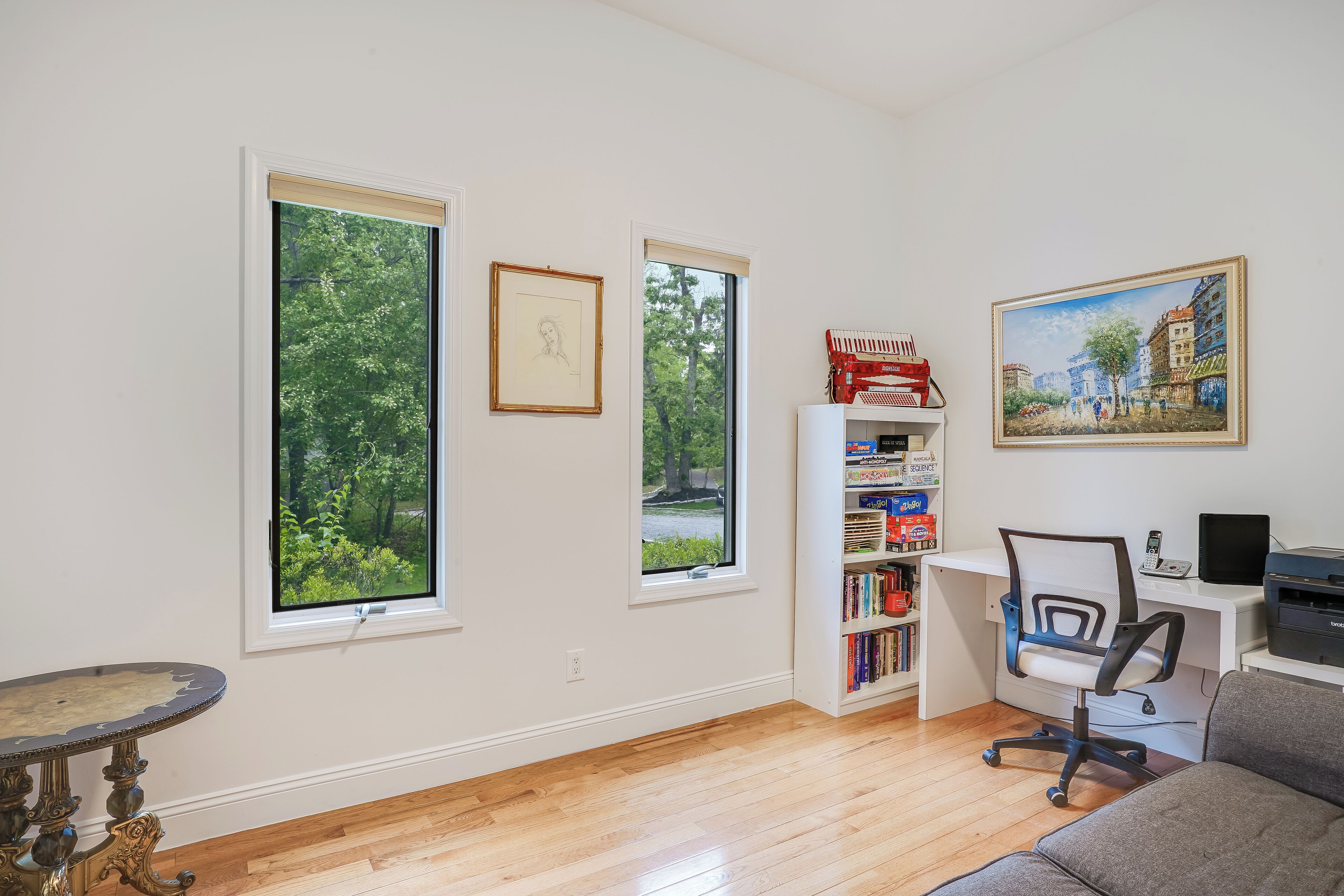 A room with large windows, a desk with a computer, a bookshelf, and a painting on the wall.
