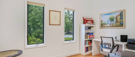 A room with large windows, a desk with a computer, a bookshelf, and a painting on the wall.