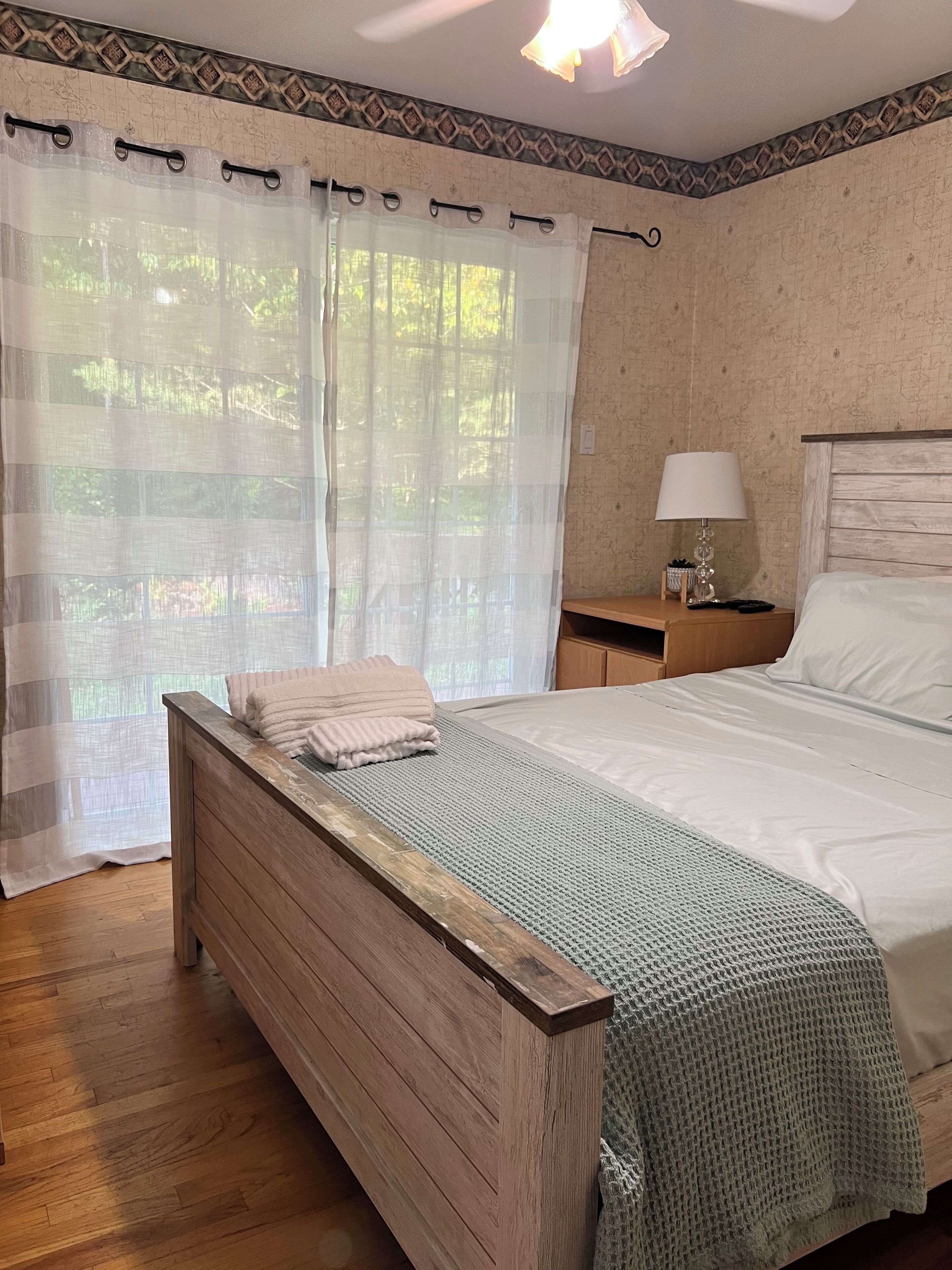 A bedroom with a wooden bed, a nightstand with a lamp, and a view of greenery through sheer curtains.