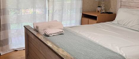 A bedroom with a wooden bed, a nightstand with a lamp, and a view of greenery through sheer curtains.