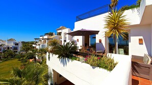 Ein weißes Gebäude mit Balkonen und einer Terrasse mit Pflanzen, vor einem strahlend blauen Himmel.