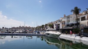 Ein Yachthafen mit vertäuten Booten, eine weiße Yacht im Vordergrund und im Hintergrund Gebäude mit Balkonen und Geschäften.