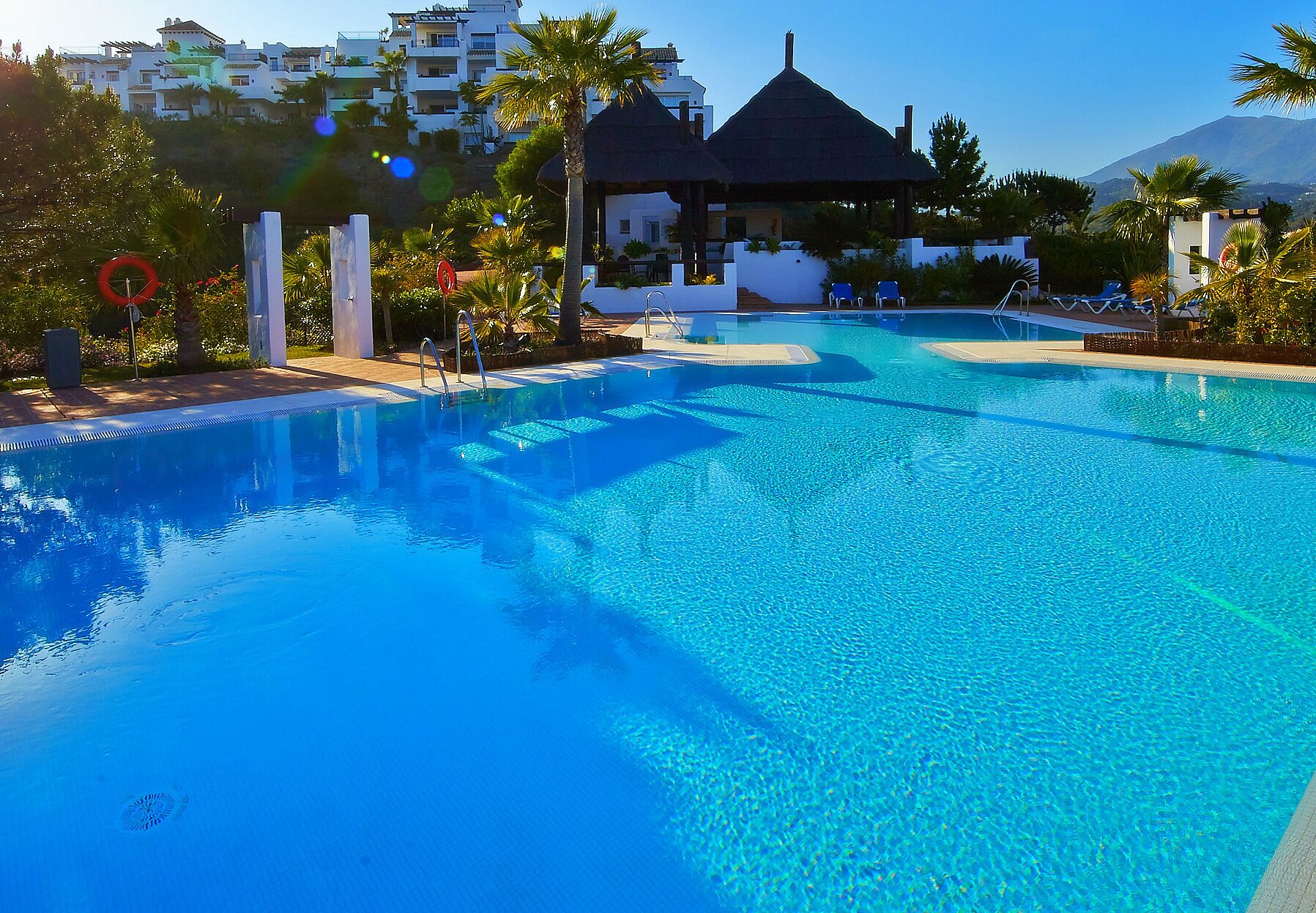 A clear blue pool with a thatched-roof gazebo, lounge chairs, and palm trees.