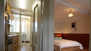 Un dormitorio con cama, cabecera de madera y mesita de noche. Un pasillo conduce a una cocina con armarios de madera y suelo de baldosas.
