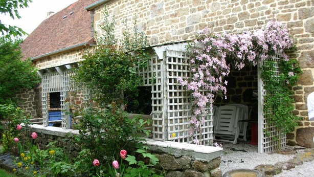 Un bâtiment en pierre avec un jardin à l’avant, comprenant un abreuvoir pour oiseaux et des plantes en fleurs.