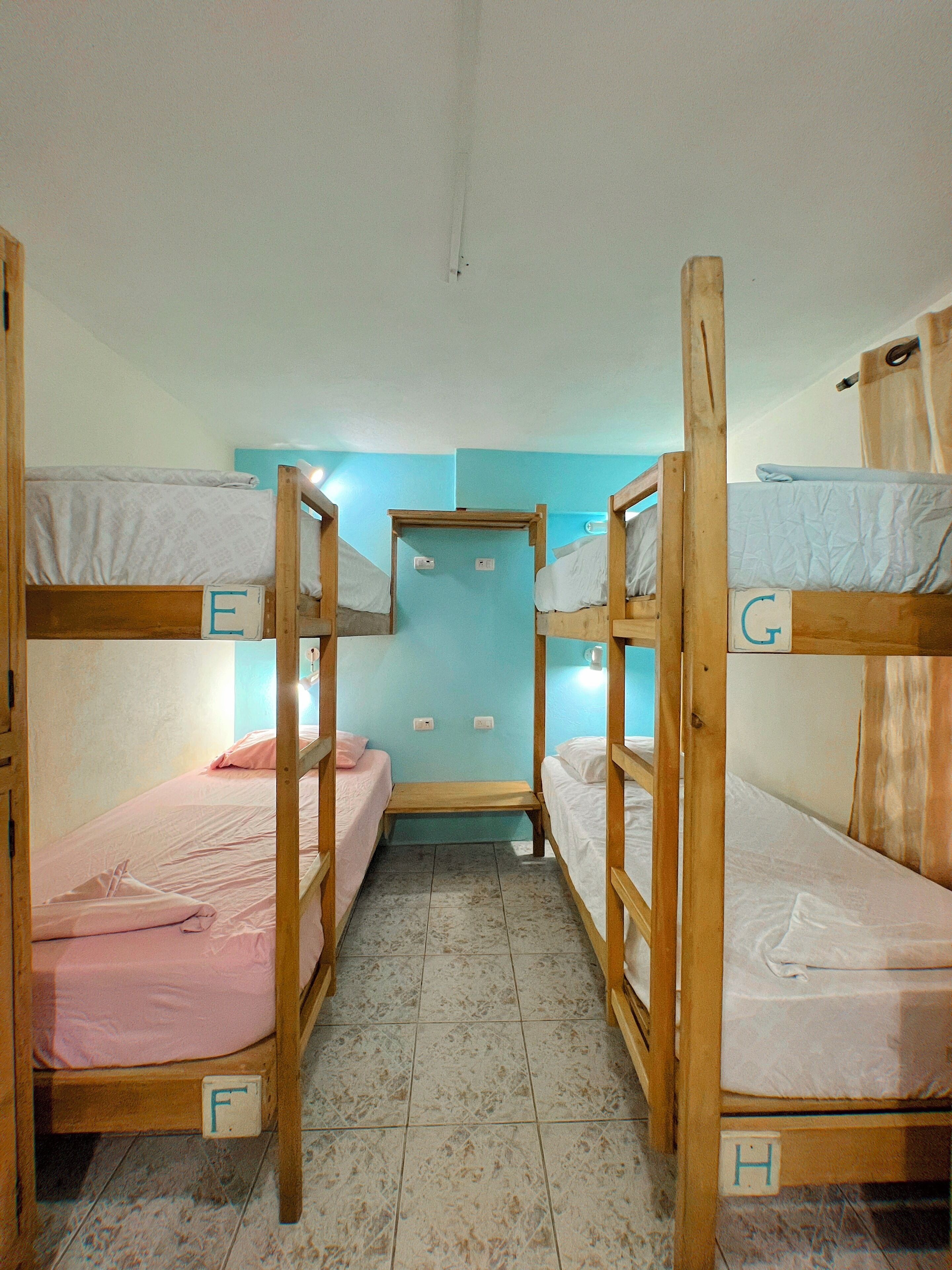 A narrow room with bunk beds labeled E, F, G, and H, a door, and a light switch on the wall.