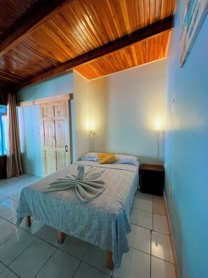 A bedroom with a wooden ceiling, a bed with a towel, two bedside lamps, and a door with a wooden panel. - Hostel Samara (Sámara)