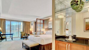Une chambre d’hôtel avec un grand lit, un bureau, une chaise, un banc, une cloison en verre et une vue sur la ville.