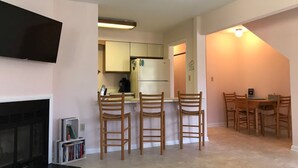 Un salon avec une cheminée, un téléviseur fixé au mur et un coin cuisine équipé d’un réfrigérateur et de placards. On y trouve trois chaises en bois et une table à manger avec des chaises.