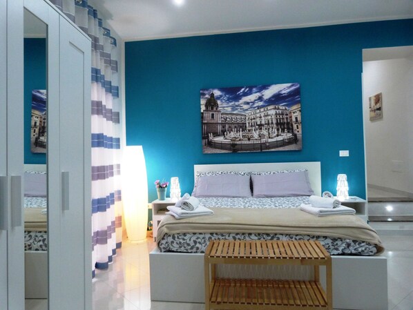 A bedroom with a large bed, a nightstand, a wooden bench, a painting of a cityscape, and a mirror reflecting the room.