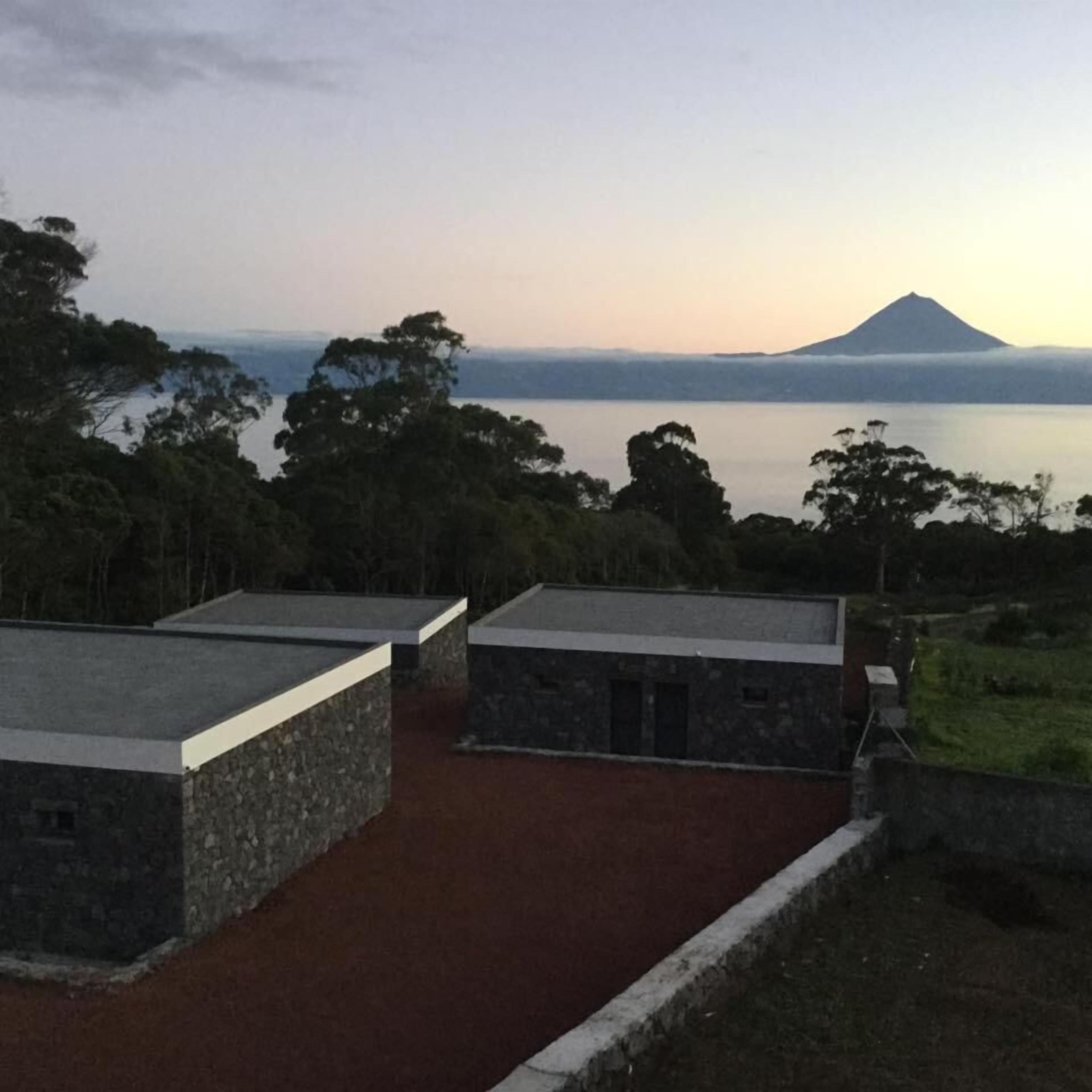 A view of a coastal area with a mountain in the background, surrounded by trees and a body of water. There are several stone buildings with flat roofs.
