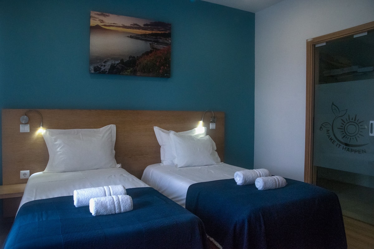 A hotel room with two beds, each with white linens and a blue bedspread. There is a painting of a sunset on the wall, and a logo on the door.