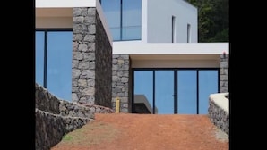 A modern two-story house with large glass windows and a stone facade, set against a mountainous backdrop. - Make It Happen Farm (Velas)