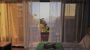 A bedroom with a bed, a bench, and a view of the outdoors through sheer curtains. - Make It Happen Farm (Velas)