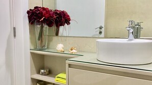 Ein Badezimmer mit einem weißen Waschbecken, einem Spiegel und einem Regal mit dekorativen Muscheln sowie einer Blumenvase.