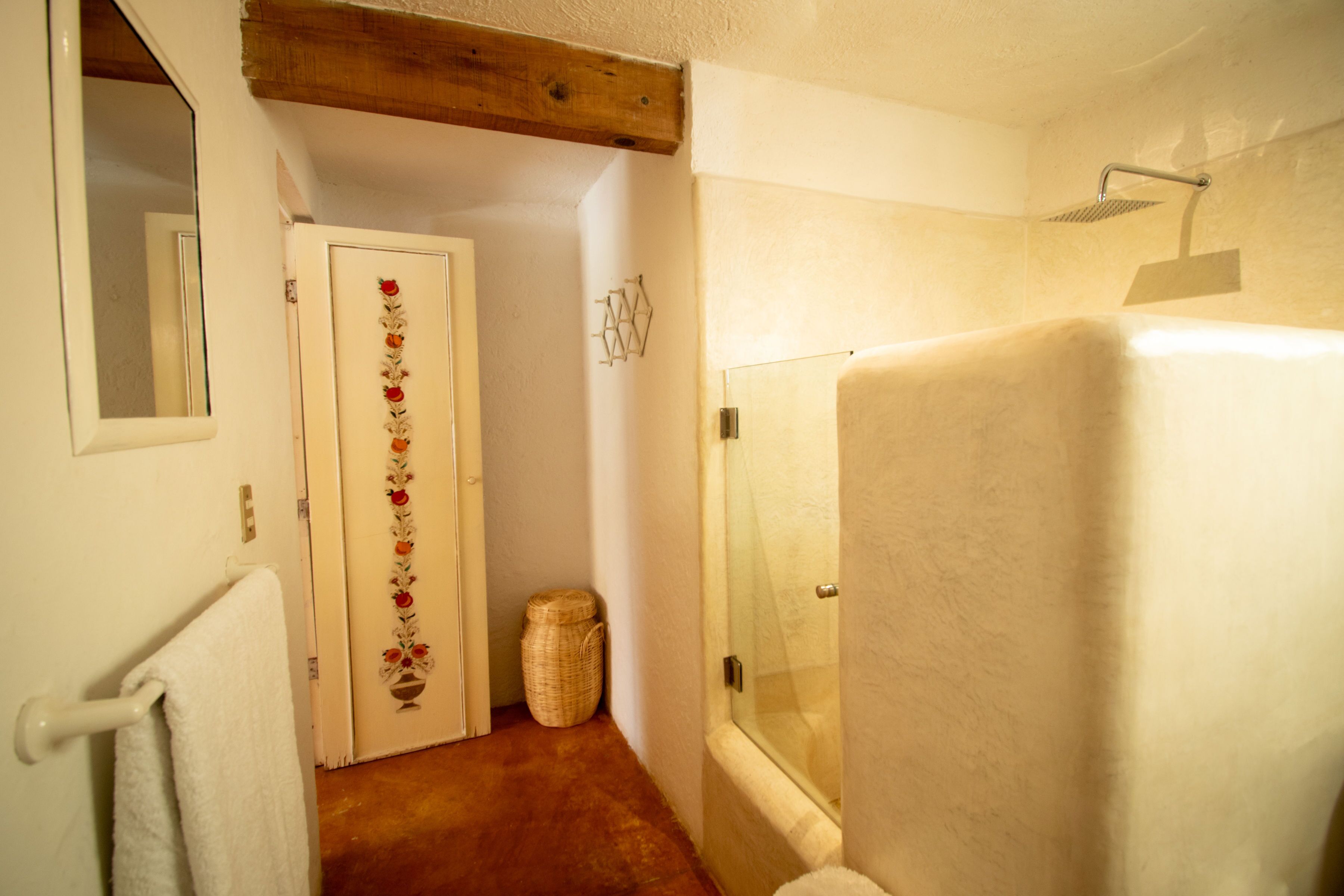 A bathroom with a bathtub, a shower, a wooden ceiling beam, a white door with a floral design, and a mirror on the wall.