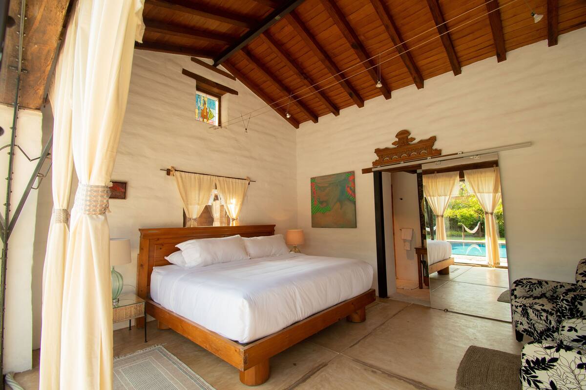A bedroom with a wooden bed, a nightstand, a lamp, and a view of an outdoor swimming pool.