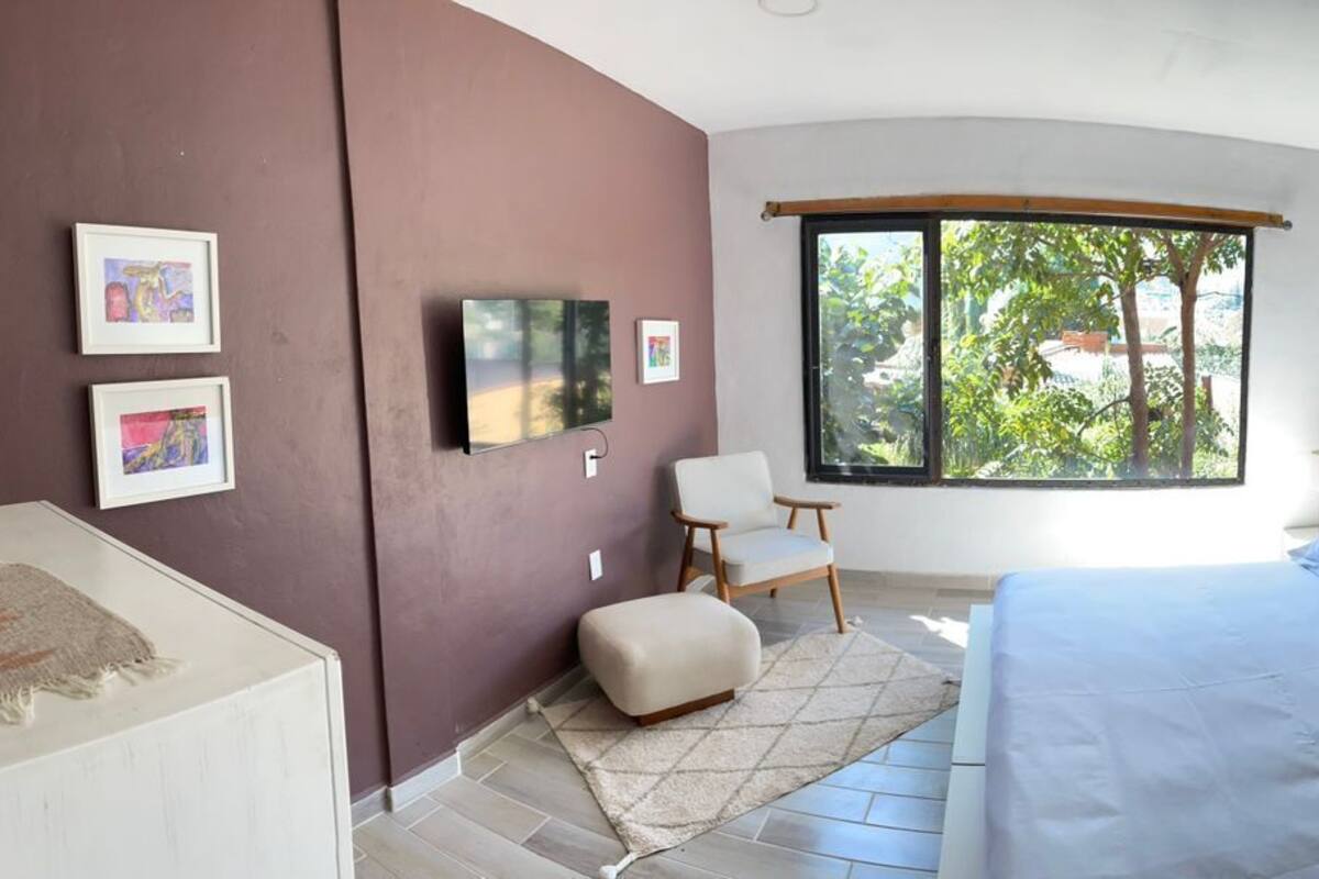 A bedroom with a bed, a chair, a window with a view of greenery, and framed pictures on the wall.