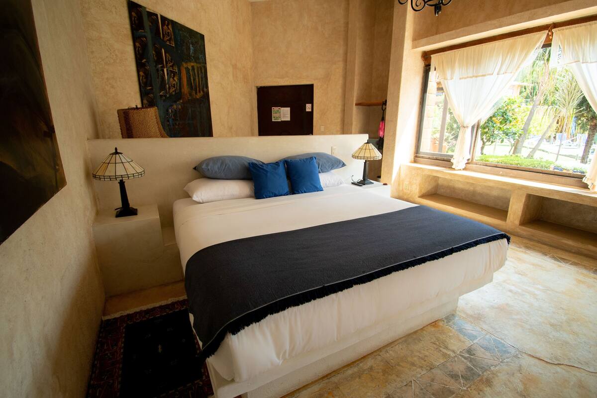 A large bed with a black bedspread, two blue pillows, and a white headboard. There is a window with sheer curtains and a view of greenery outside.